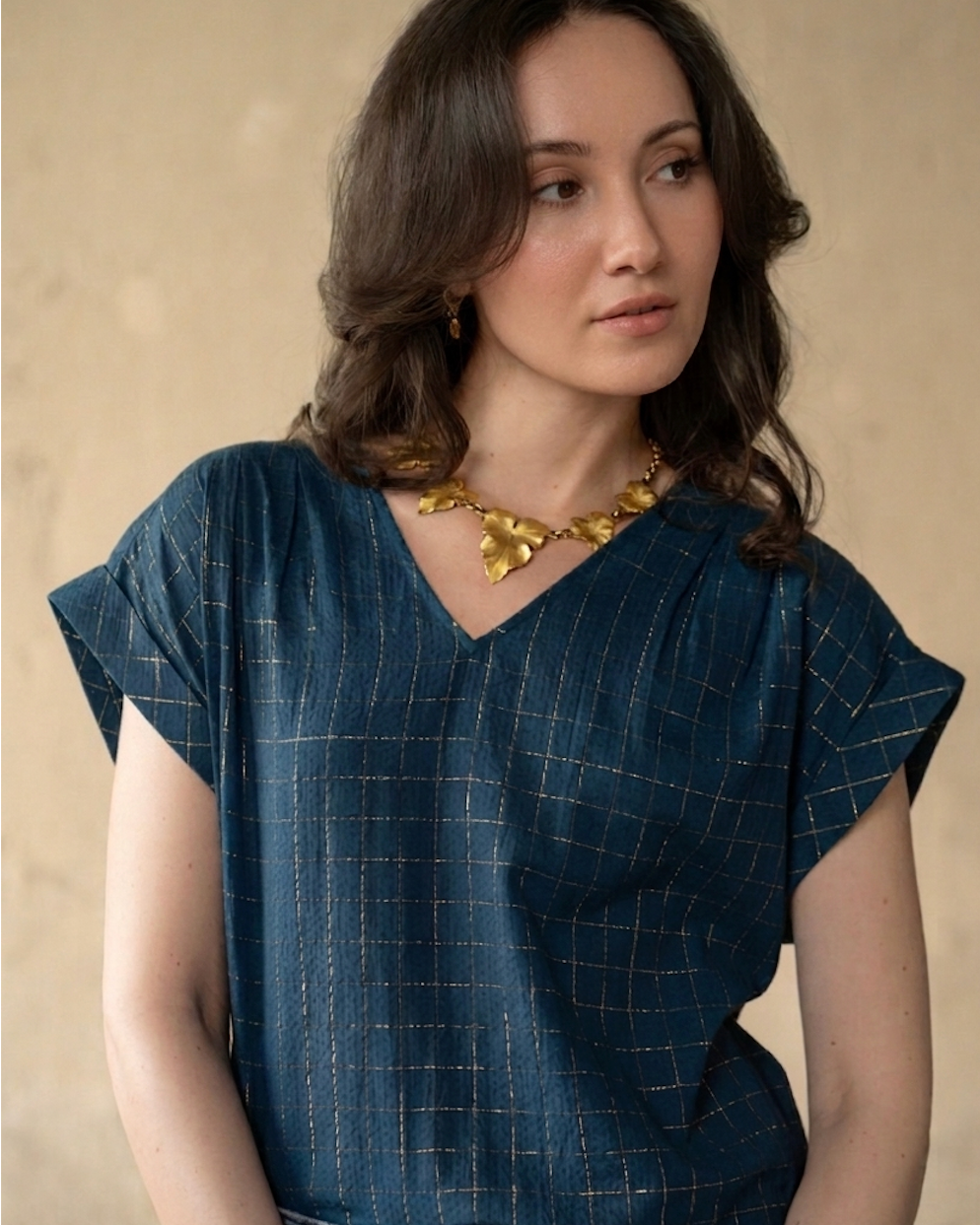 A woman wearing a dark blue V-neck top with a subtle gold grid pattern, styled with a chunky gold necklace and earrings, set against a neutral beige background.