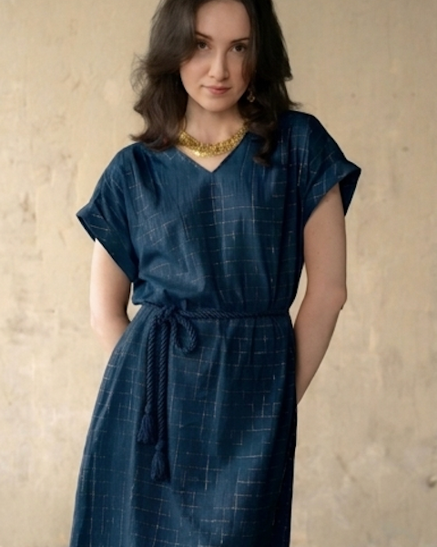 A woman wearing a dark blue V-neck dress with a subtle grid pattern and a braided tie belt at the waist, standing against a textured beige wall.