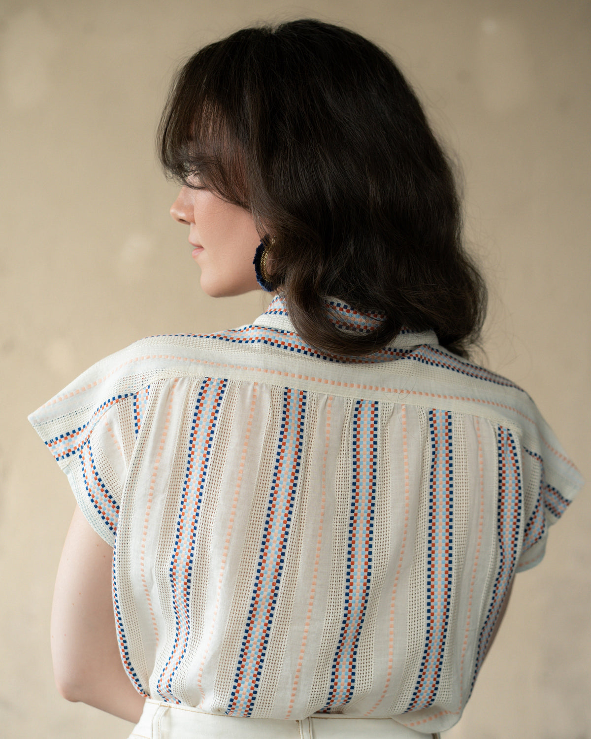 A woman shown from the back wearing a cream-colored textured blouse with vertical stripes in blue, orange, and red, paired with white pants against a neutral beige background.
