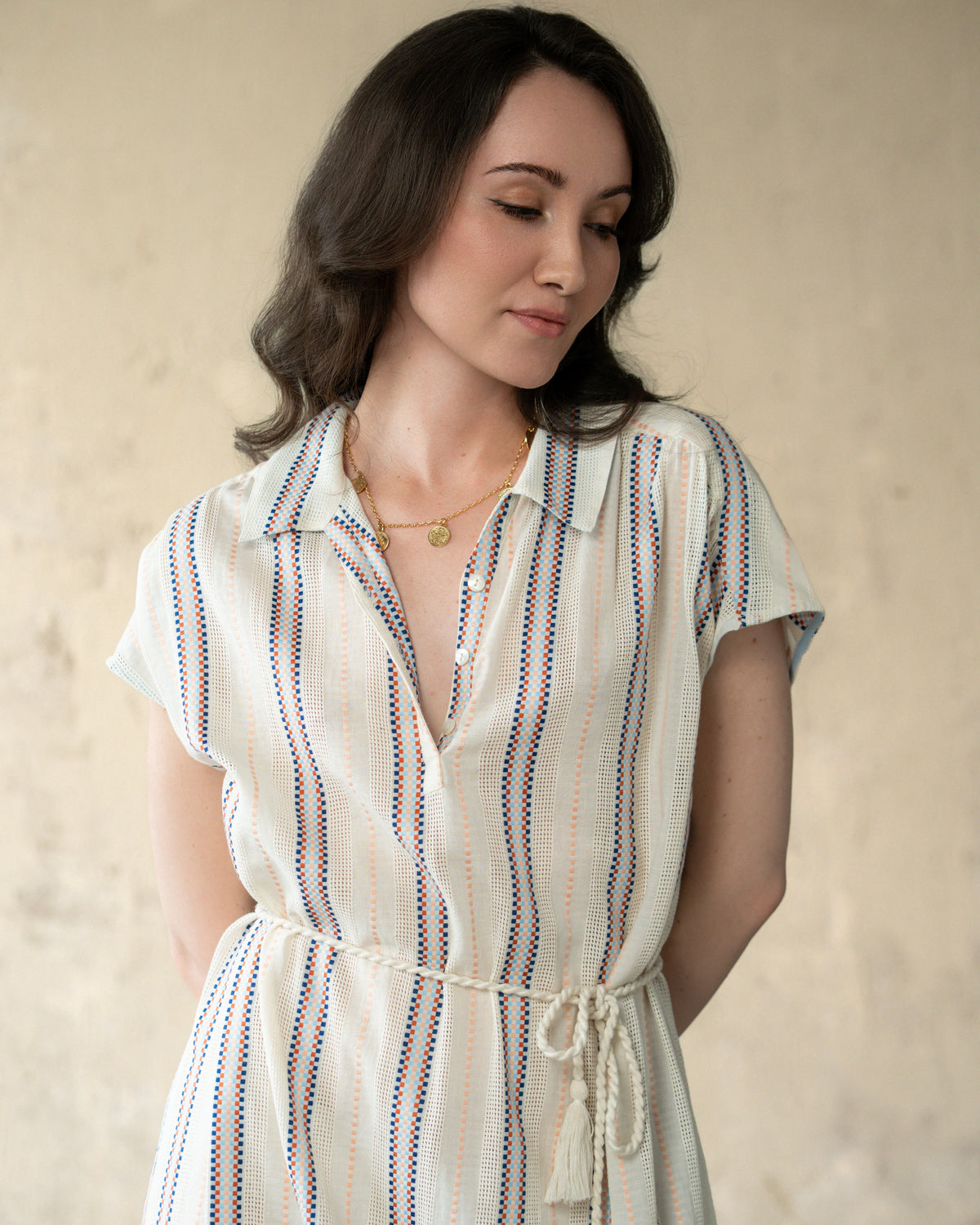 A woman wearing a short-sleeve collared shirt dress with vertical blue and orange stripes on a cream base, featuring a tie waist and tassel, set against a textured beige wall.