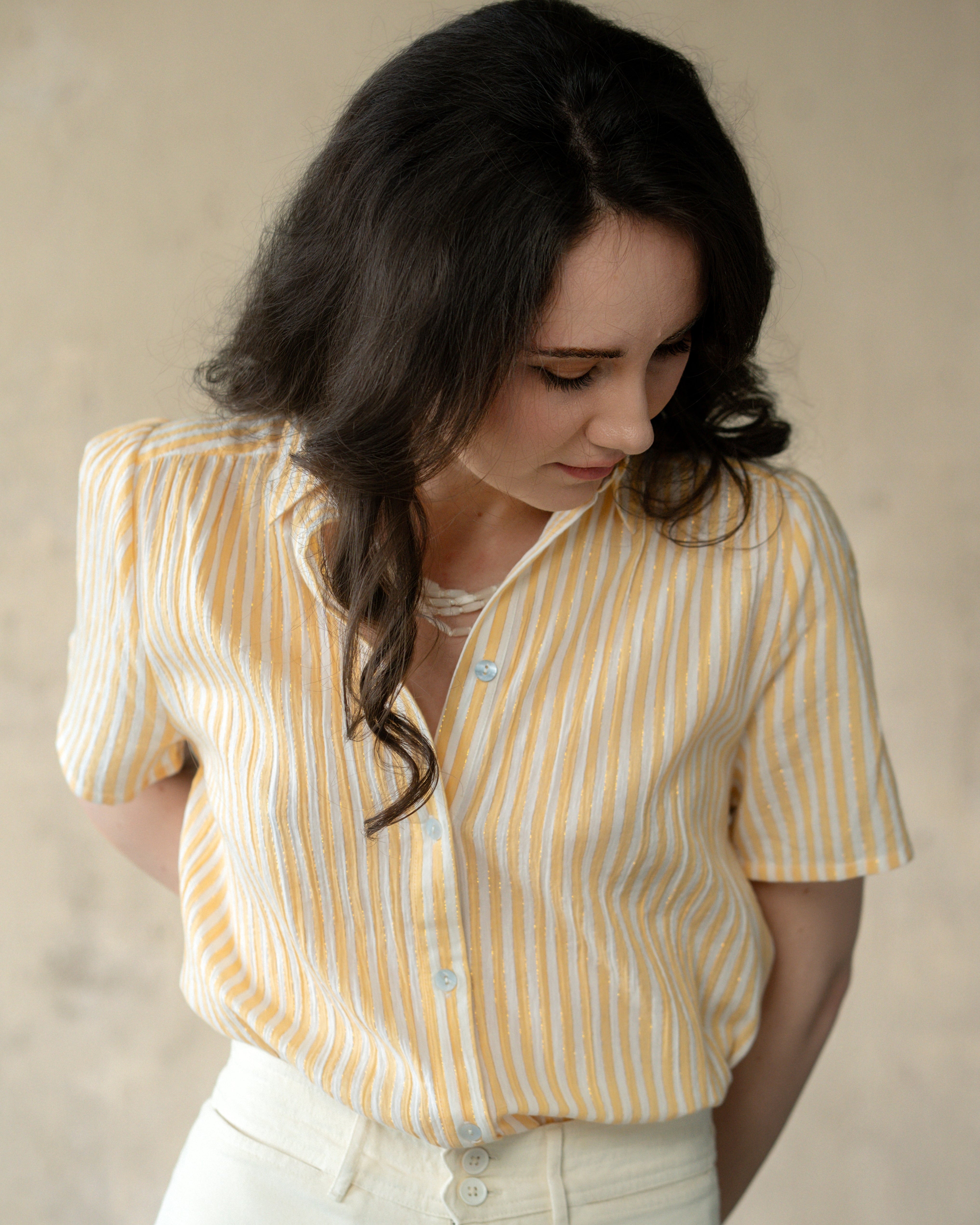 A woman wearing a short-sleeve button-up shirt with vertical yellow and white stripes, paired with off-white pants, set against a neutral beige background.