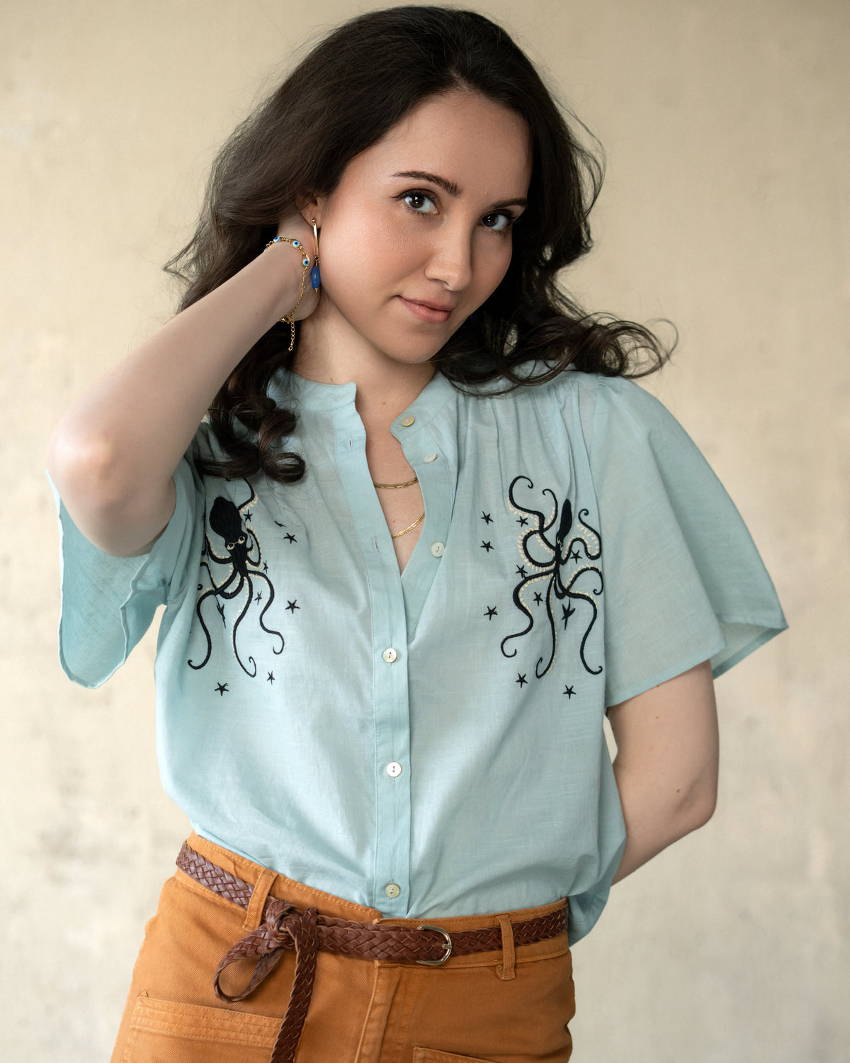 A woman wearing a light blue short-sleeve button-up shirt with black octopus and star embroidery, paired with brown pants and a braided belt, posing against a neutral beige wall.