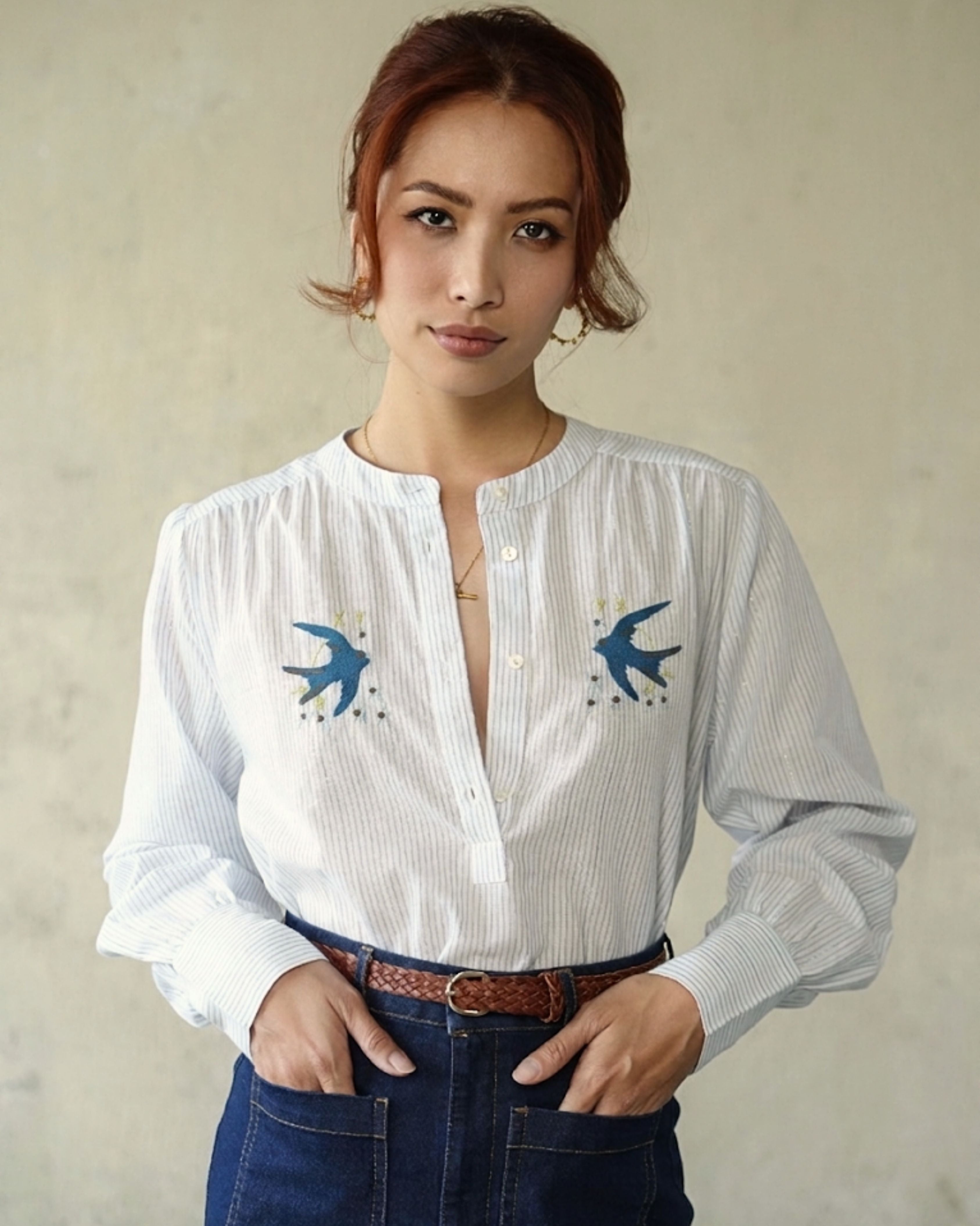 A woman wearing a white long-sleeve button-up shirt with blue swallow bird embroidery on the chest, paired with dark blue jeans and a brown belt, standing against a textured beige wall.