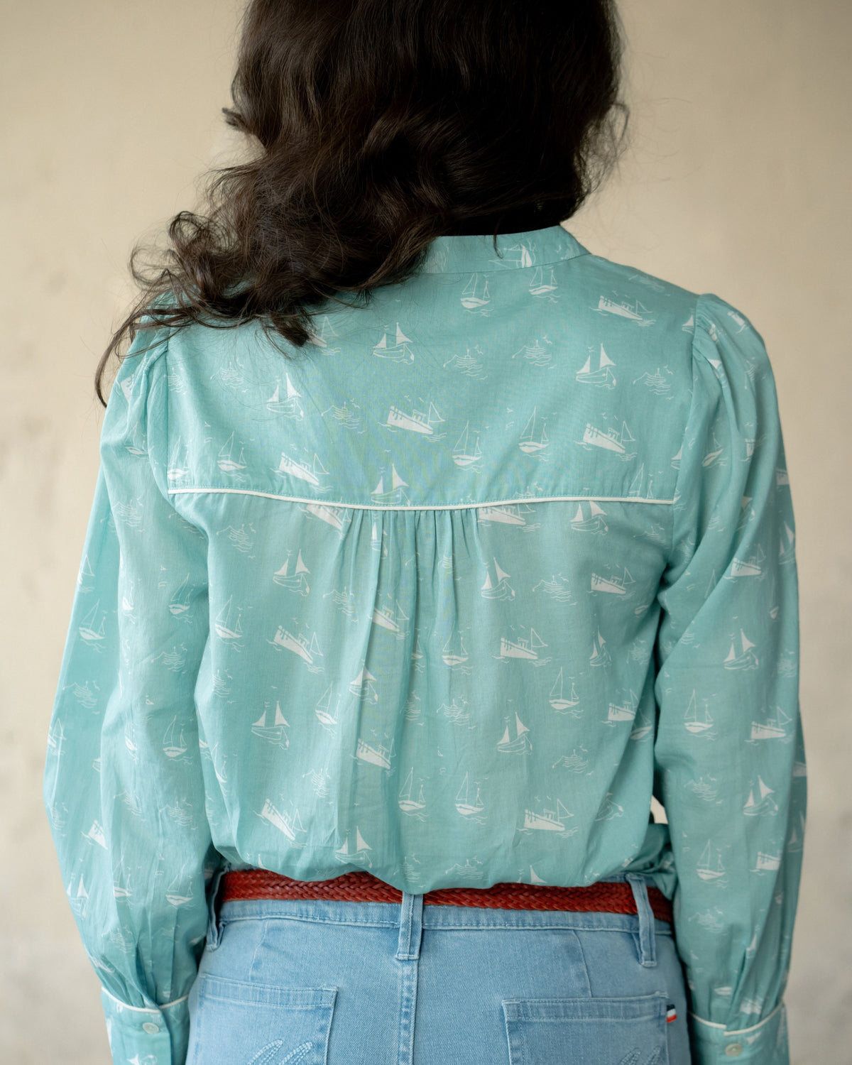 A female model shown from the back wearing a light aqua long-sleeve blouse with a white sailboat print and a red belt, paired with light blue jeans against a neutral beige background.