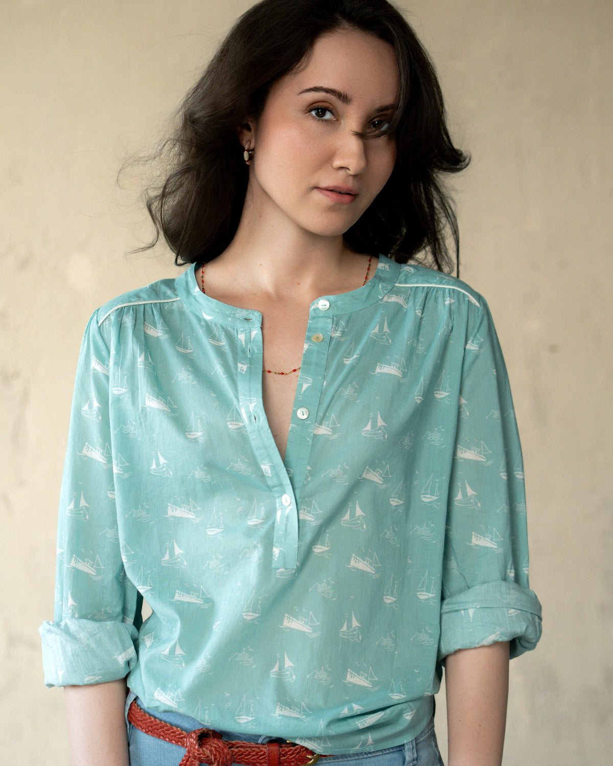 A woman wearing a light aqua blue long-sleeve henley shirt with a white sailboat pattern, paired with light blue jeans and a red braided belt, standing against a neutral beige wall.