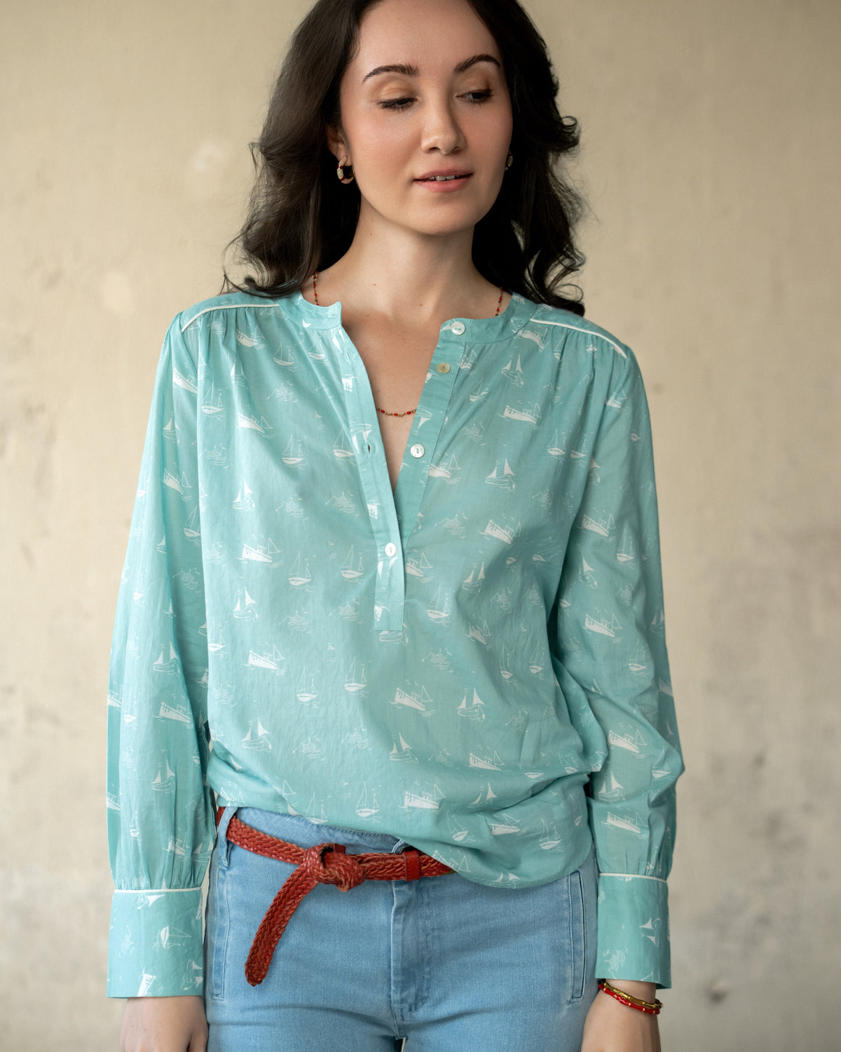 A woman wearing a light teal long-sleeve blouse with a white sailboat print, tucked into light blue jeans with a red belt tied at the waist, standing against a textured beige wall.
