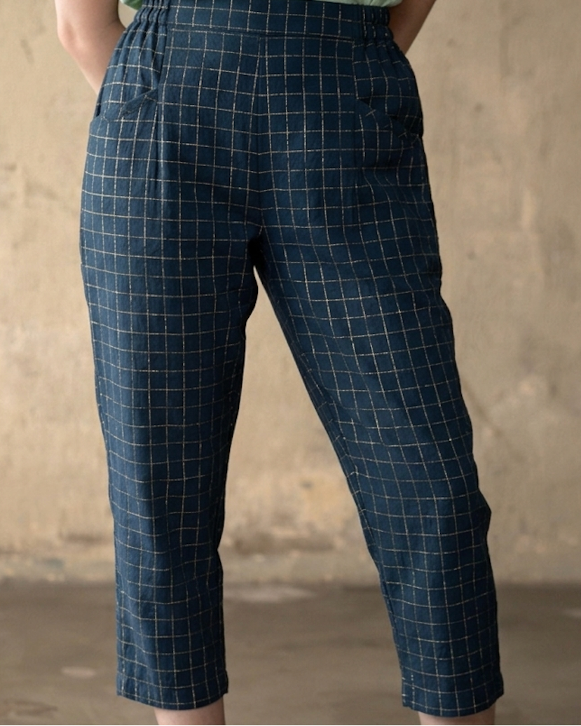 A female model wearing dark navy cropped pants with a fine light grid pattern, paired with a light green top, standing against a textured beige wall.