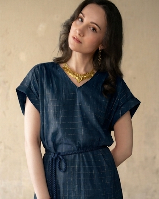 A woman wearing a dark navy blue textured dress with a tie waist and short sleeves, styled with a chunky gold necklace and earrings, standing against a neutral beige wall.