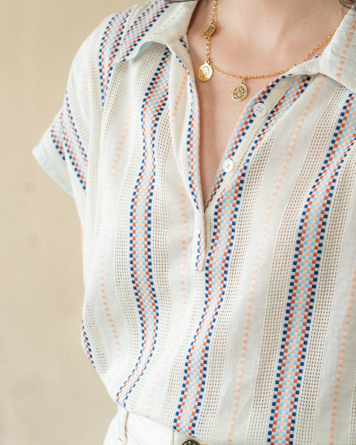 A close-up of a person wearing a white textured short-sleeve button-up shirt with vertical stripes in blue and orange, paired with a gold necklace.