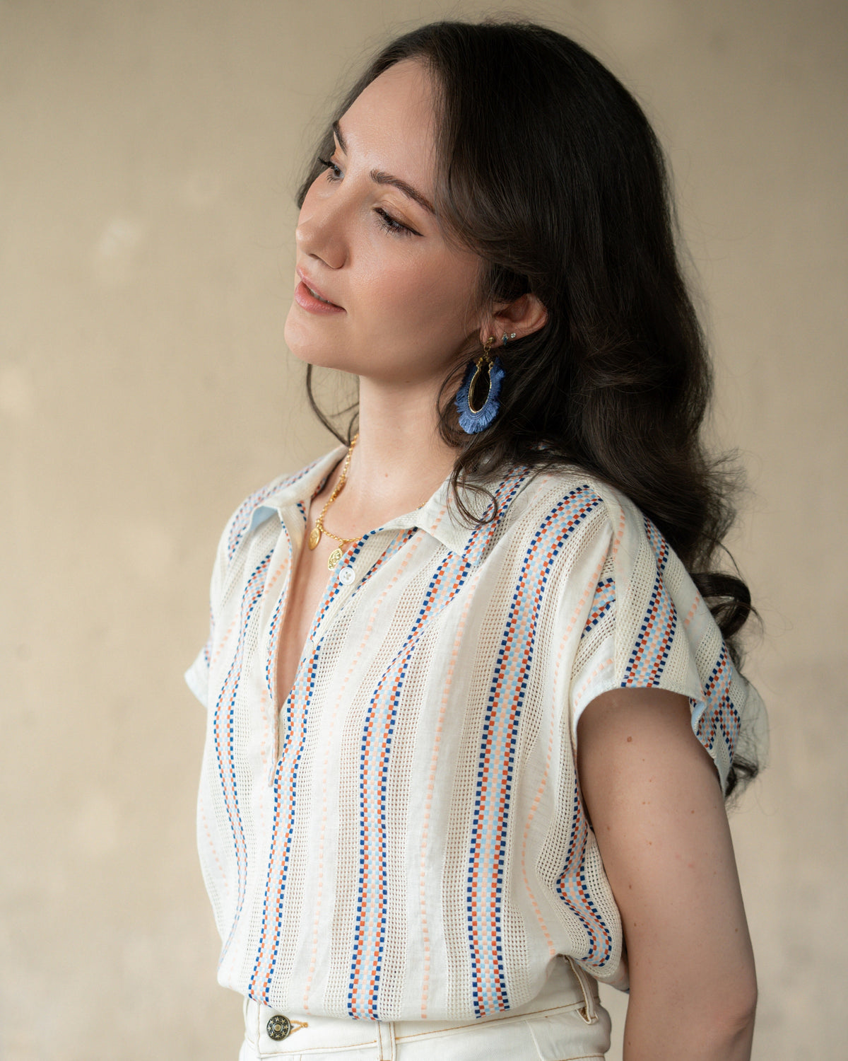A woman wearing a short-sleeve white textured blouse with vertical blue and red striped patterns, paired with white pants and blue hoop earrings, set against a neutral beige background.
