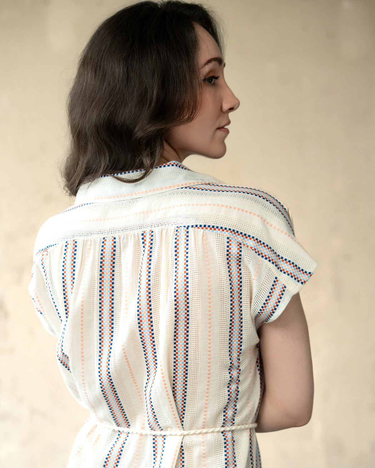 A woman shown from the back wearing a white textured short-sleeve top with vertical blue and orange dotted stripes, set against a plain beige background.