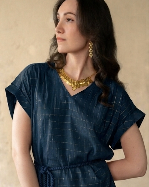 A woman wearing a dark navy blue textured dress with a tie waist and gold jewelry, posing against a neutral beige background.