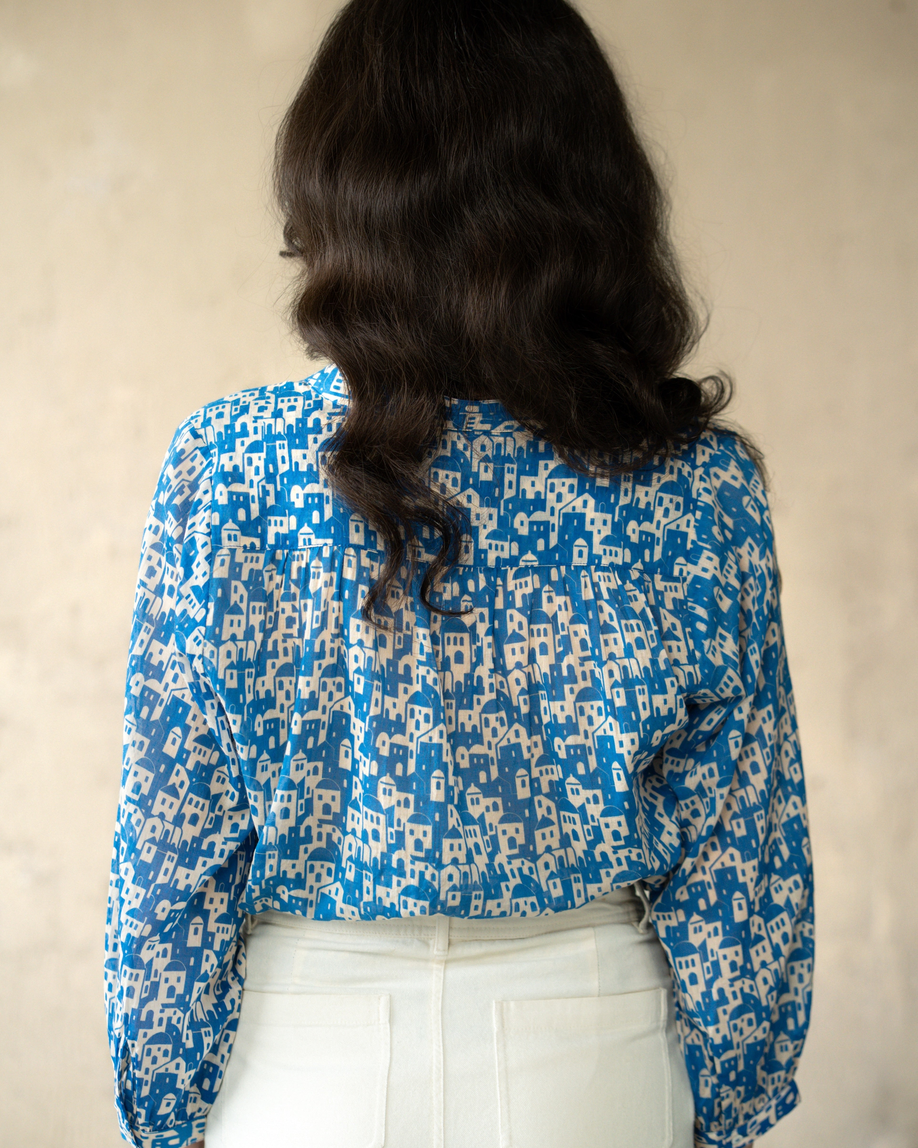 A woman viewed from behind wearing a blue and white patterned blouse with long sleeves and white pants, standing against a textured beige wall.