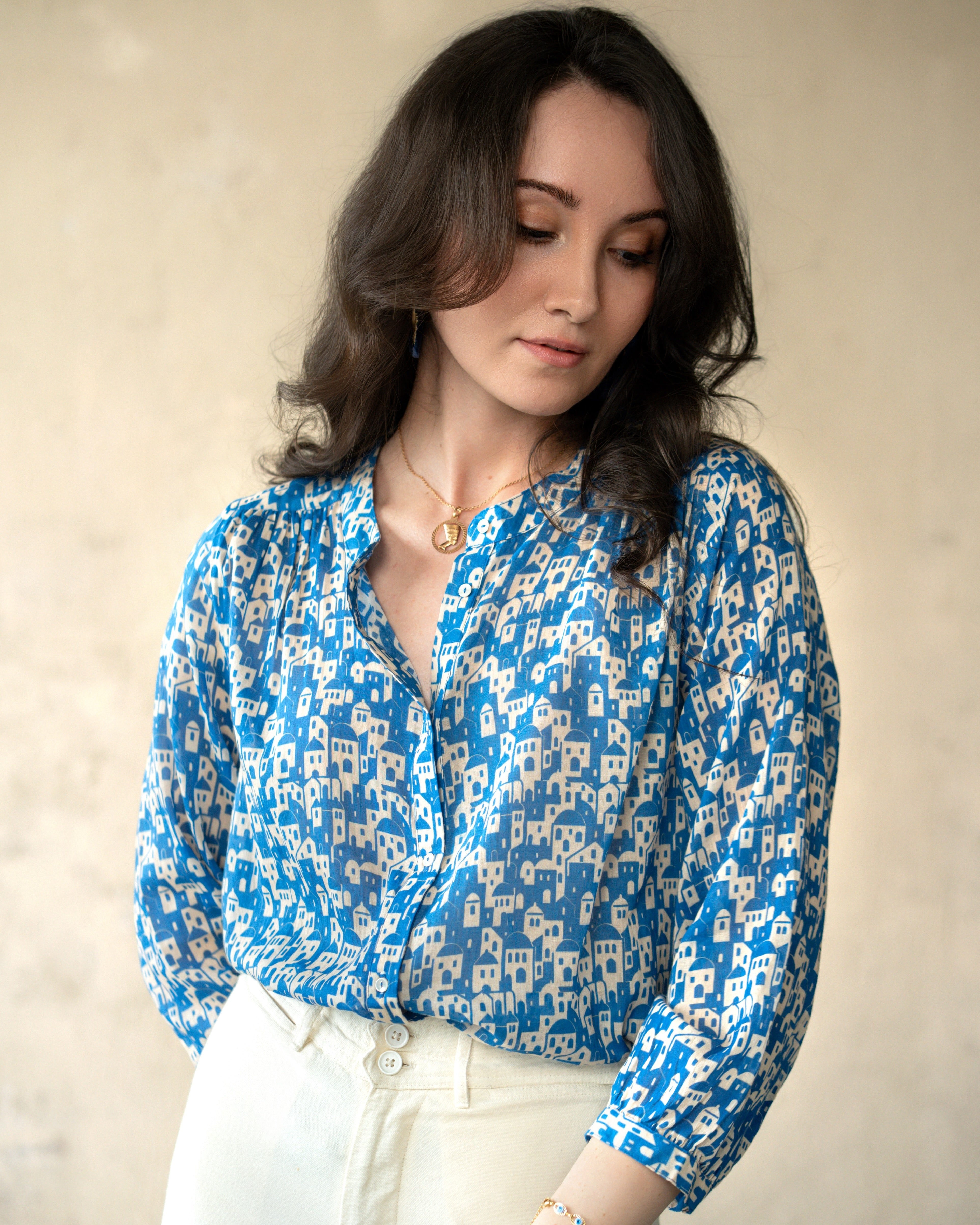 A woman wearing a blue and white patterned blouse with white pants, standing against a neutral beige wall.