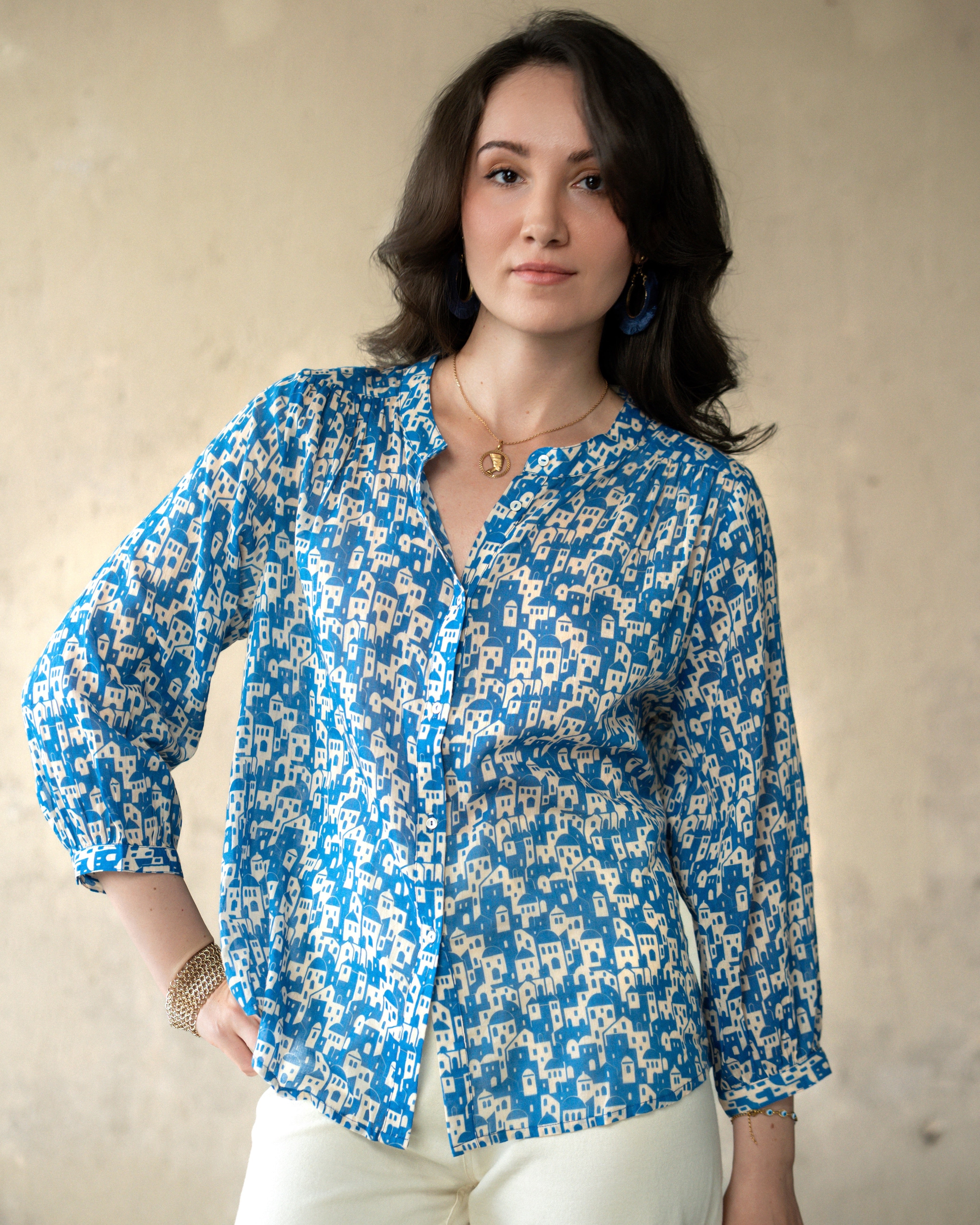 A woman wearing a blue and white patterned long-sleeve blouse with a V-neck and button front, paired with cream pants, standing against a textured beige wall.