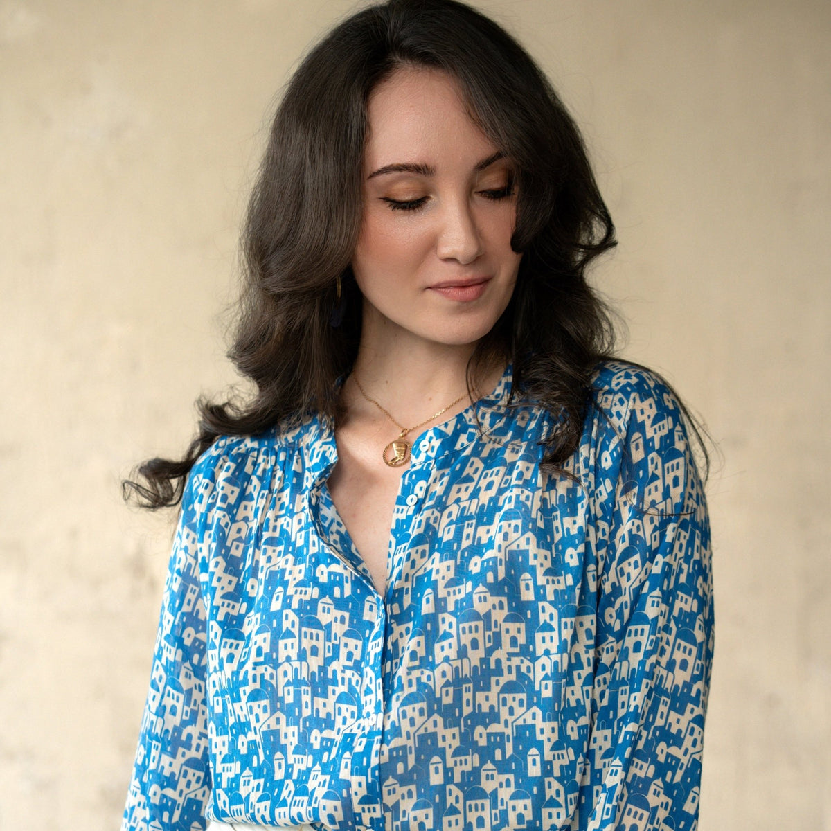 A woman wearing a blue and cream patterned long-sleeve blouse tucked into cream trousers, standing against a textured beige wall.