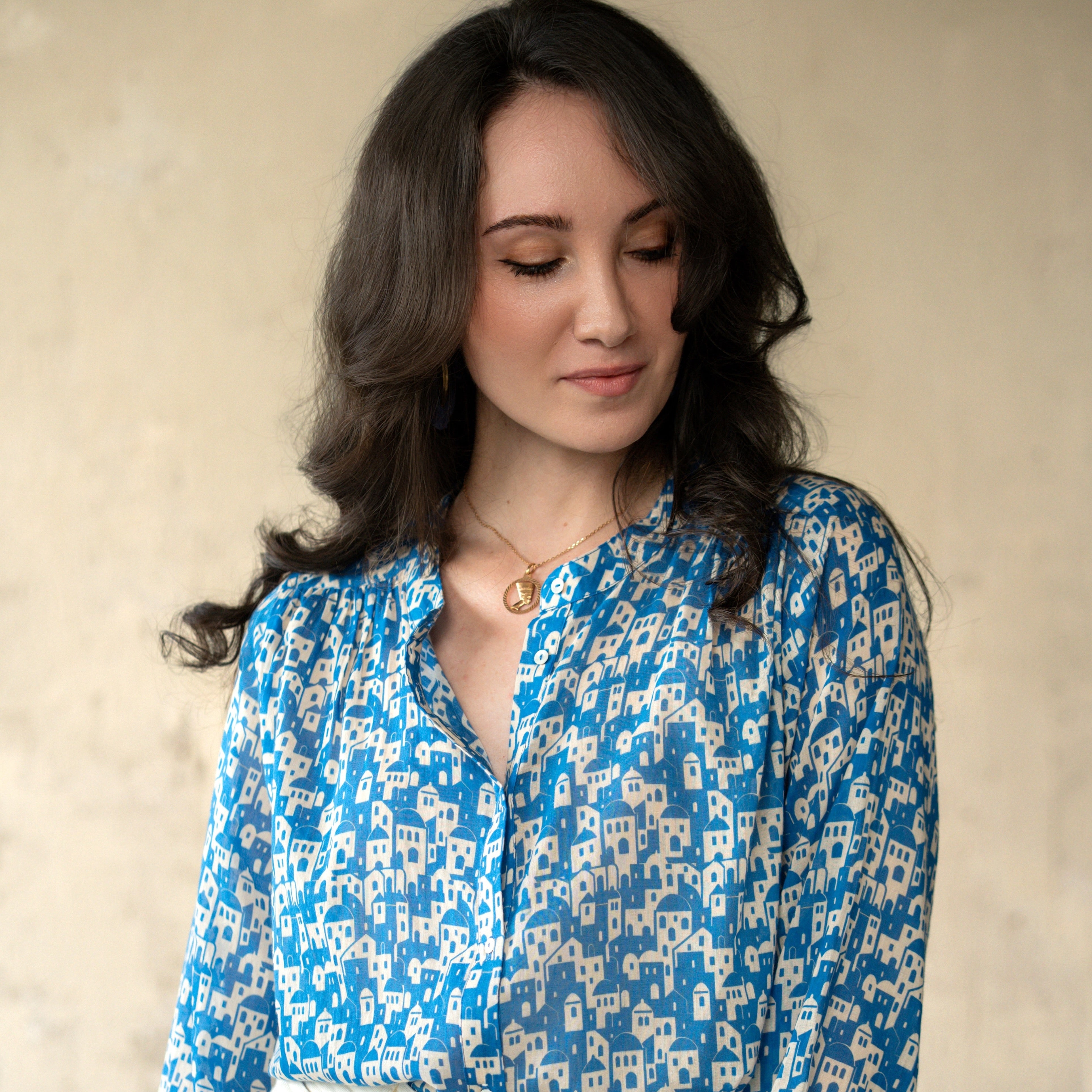 A woman wearing a blue and cream patterned long-sleeve blouse tucked into cream trousers, standing against a textured beige wall.