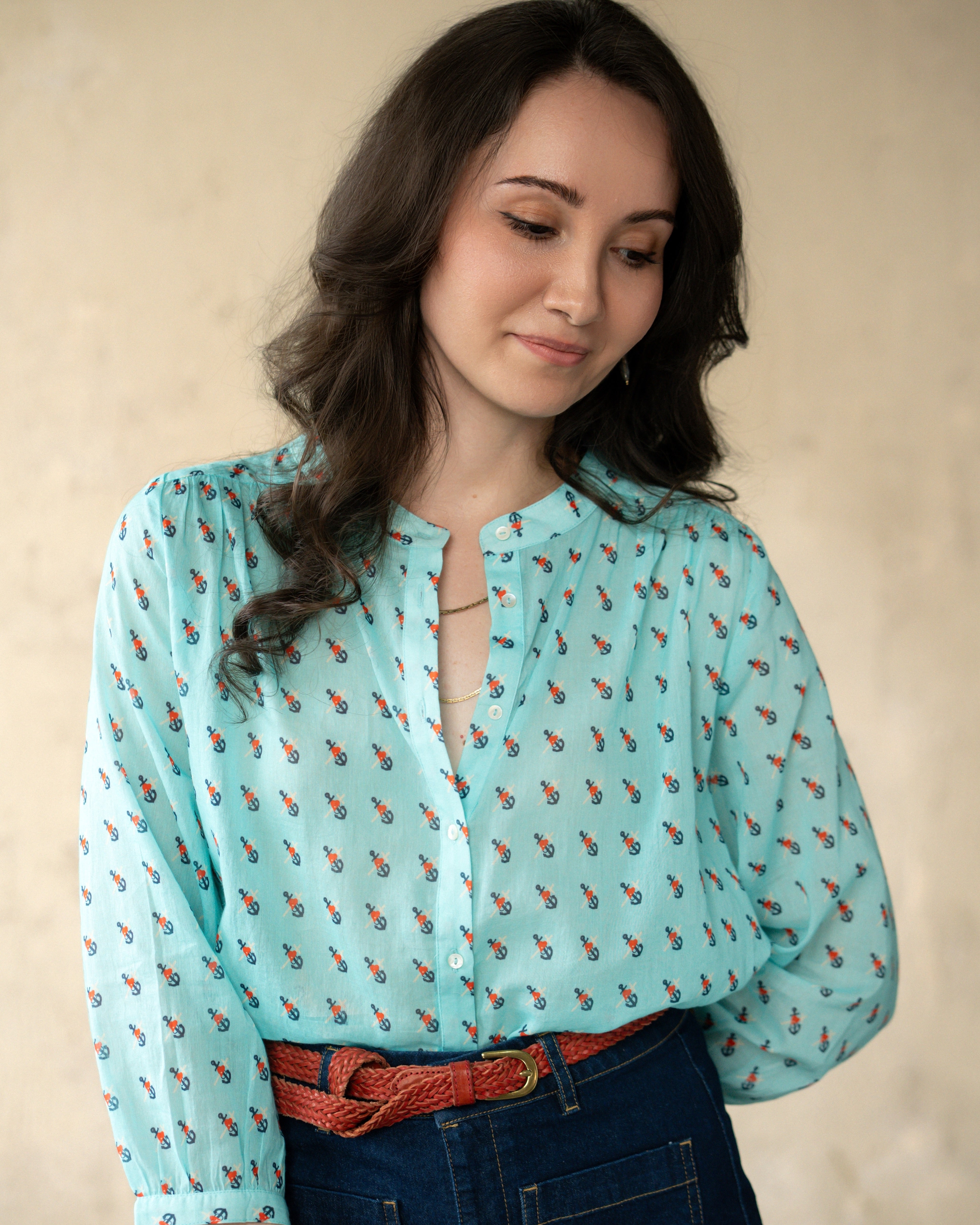 A woman wearing a light blue long-sleeve blouse with a small red and green anchor pattern, paired with a red woven belt and dark blue jeans, set against a neutral beige background.