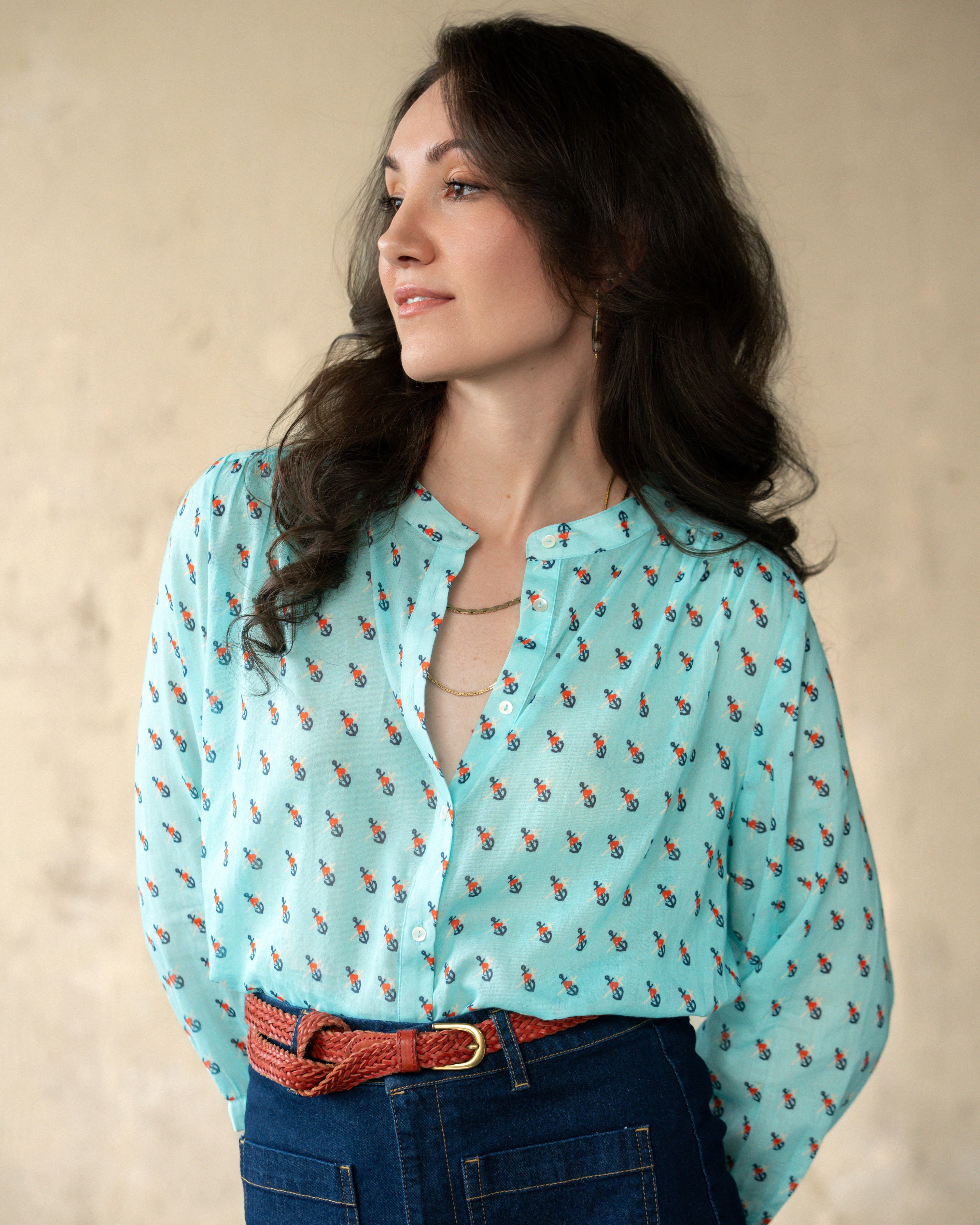 A woman wearing a light blue long-sleeve button-up blouse with a small red and orange anchor print, paired with a red woven belt and dark blue jeans, posing against a textured beige background.