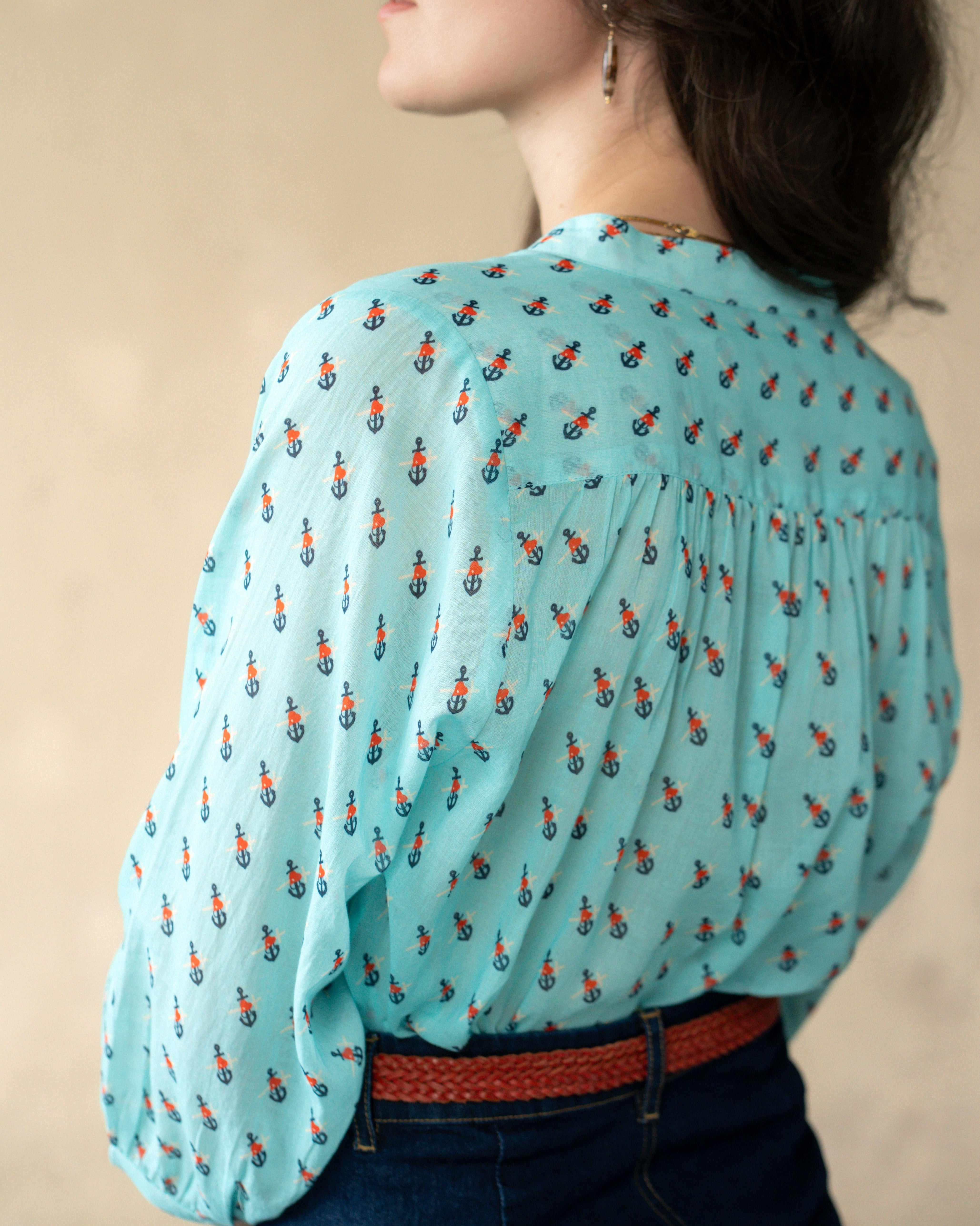A woman shown from the back wearing a light aqua blue blouse with a small repeating orange and black anchor print, paired with dark blue jeans and a brown belt.