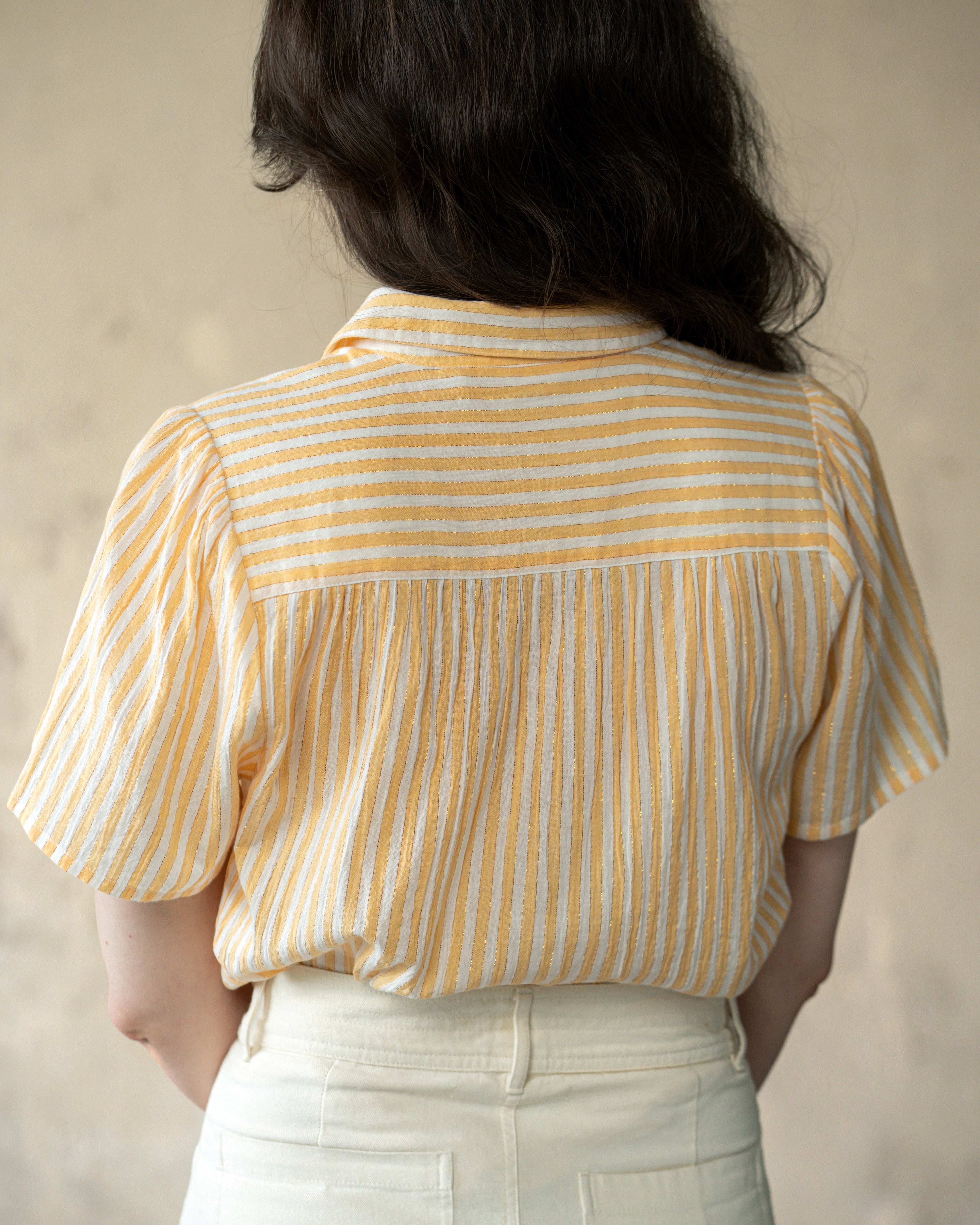 A person shown from the back wearing a yellow and white vertically striped short-sleeve blouse tucked into cream-colored pants, set against a neutral textured beige background.