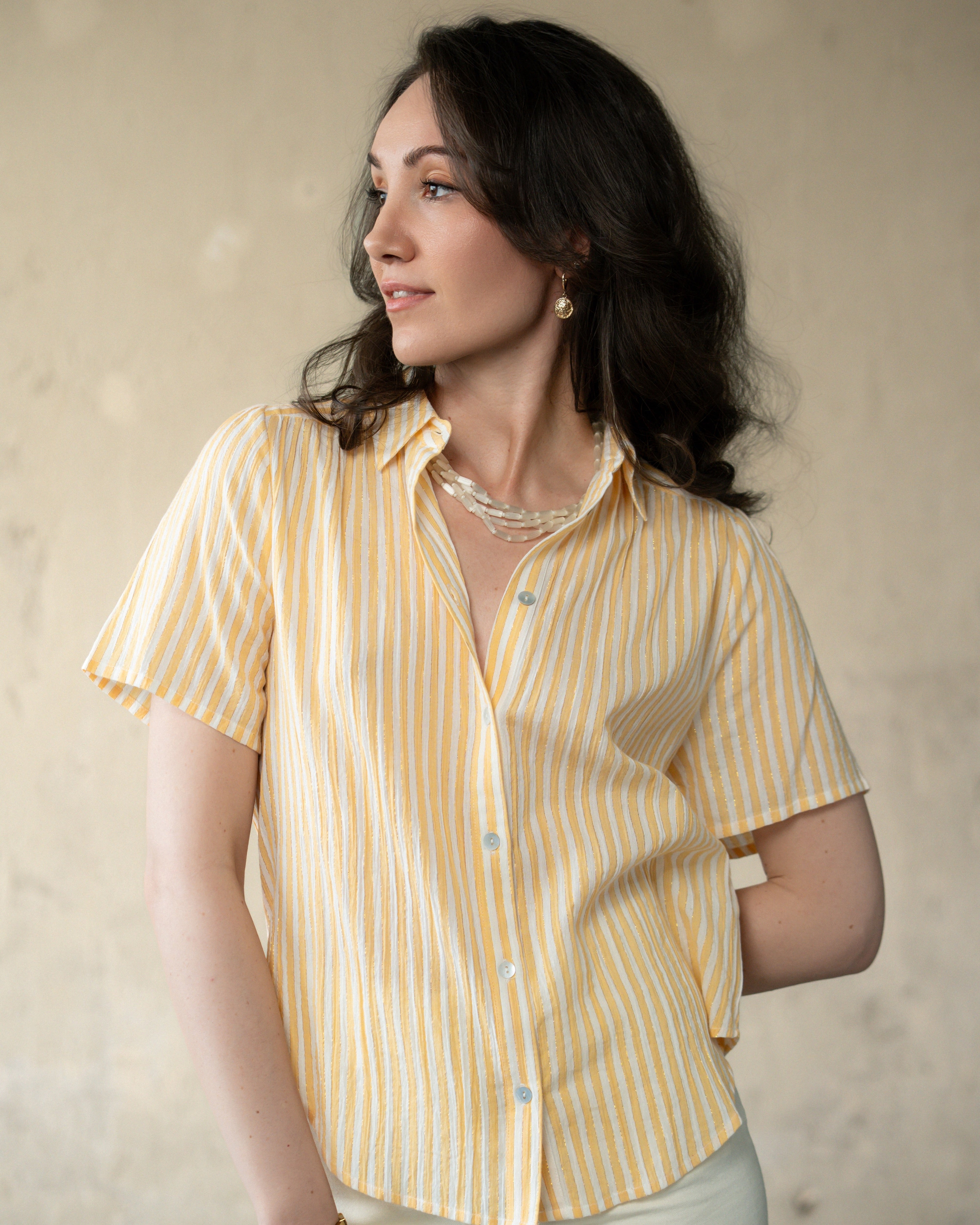 A woman wearing a short-sleeve button-up shirt with vertical yellow and white stripes, paired with light-colored pants, set against a neutral textured beige background.