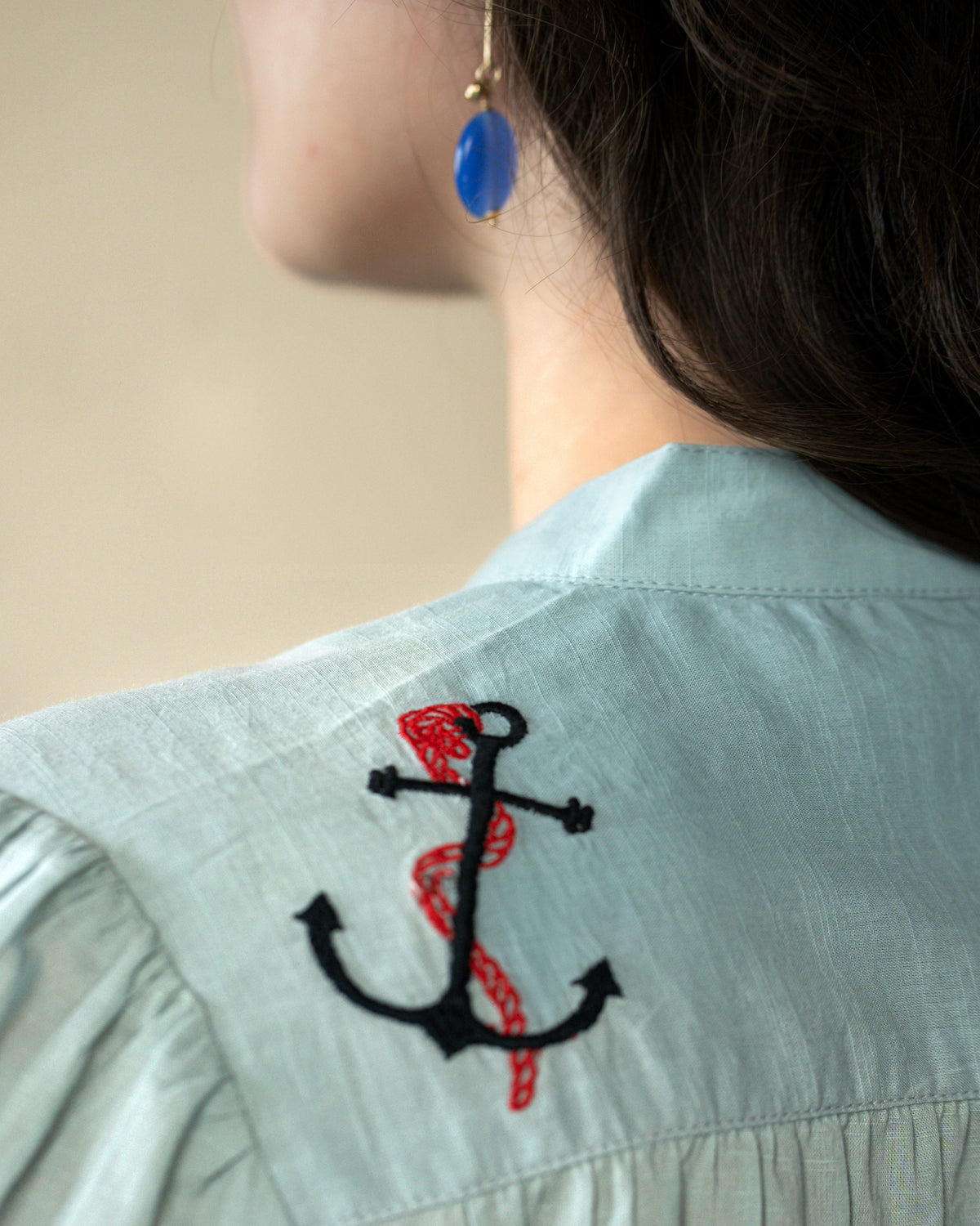 Close-up of a person wearing a pale blue shirt with an embroidered black anchor and red snake motif on the shoulder, with a blue teardrop earring visible.
