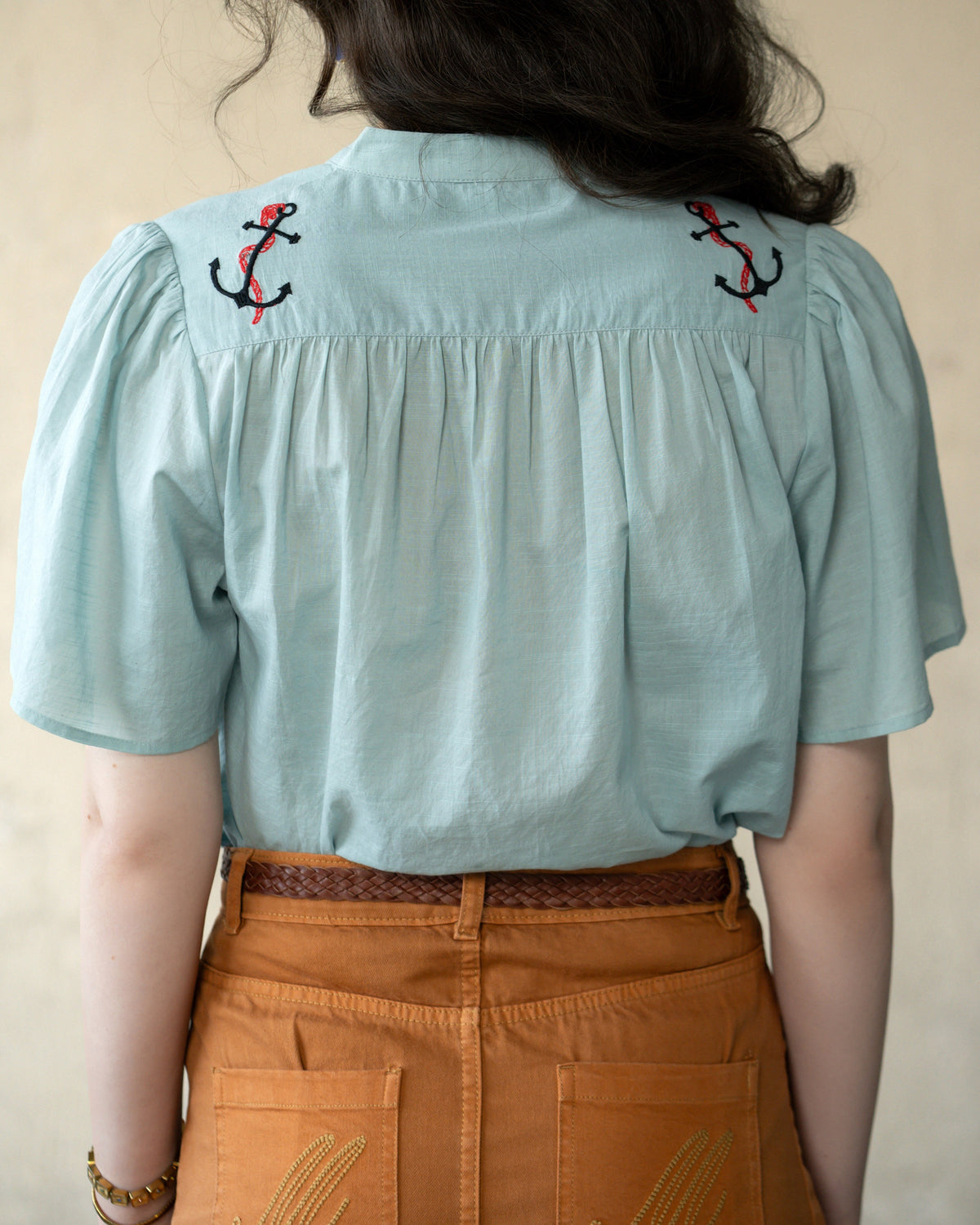 A female model is shown from the back wearing a light blue short-sleeve blouse with embroidered anchor motifs near the shoulders and brown pants.