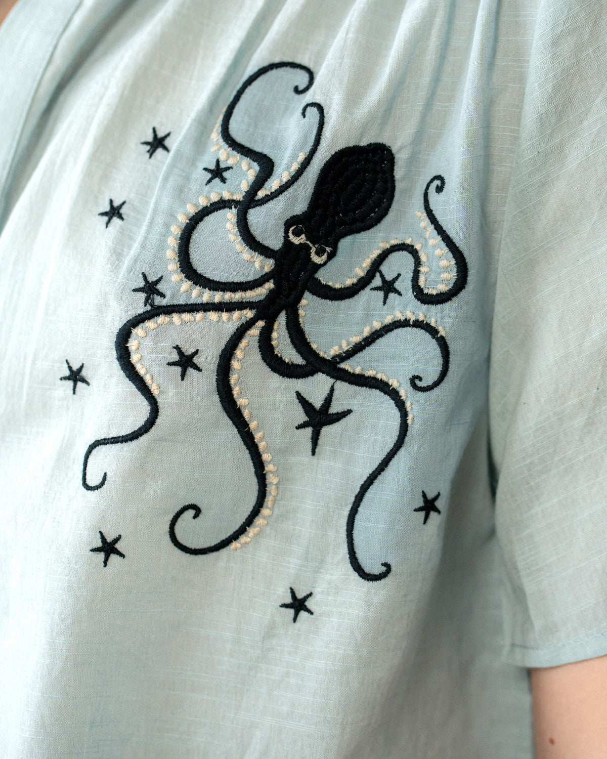 A close-up of a light sage green linen top featuring a black embroidered octopus and star pattern with a delicate gold beaded outline.