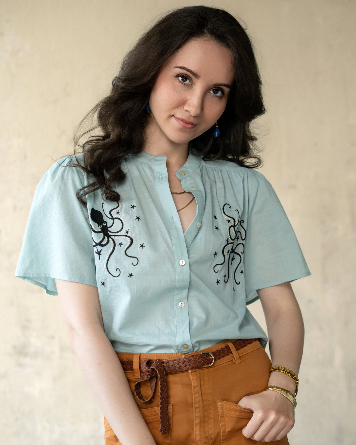 A woman wearing a light blue short-sleeve button-up shirt with black octopus and star embroidery, paired with brown pants and a brown belt, posing against a textured beige wall.