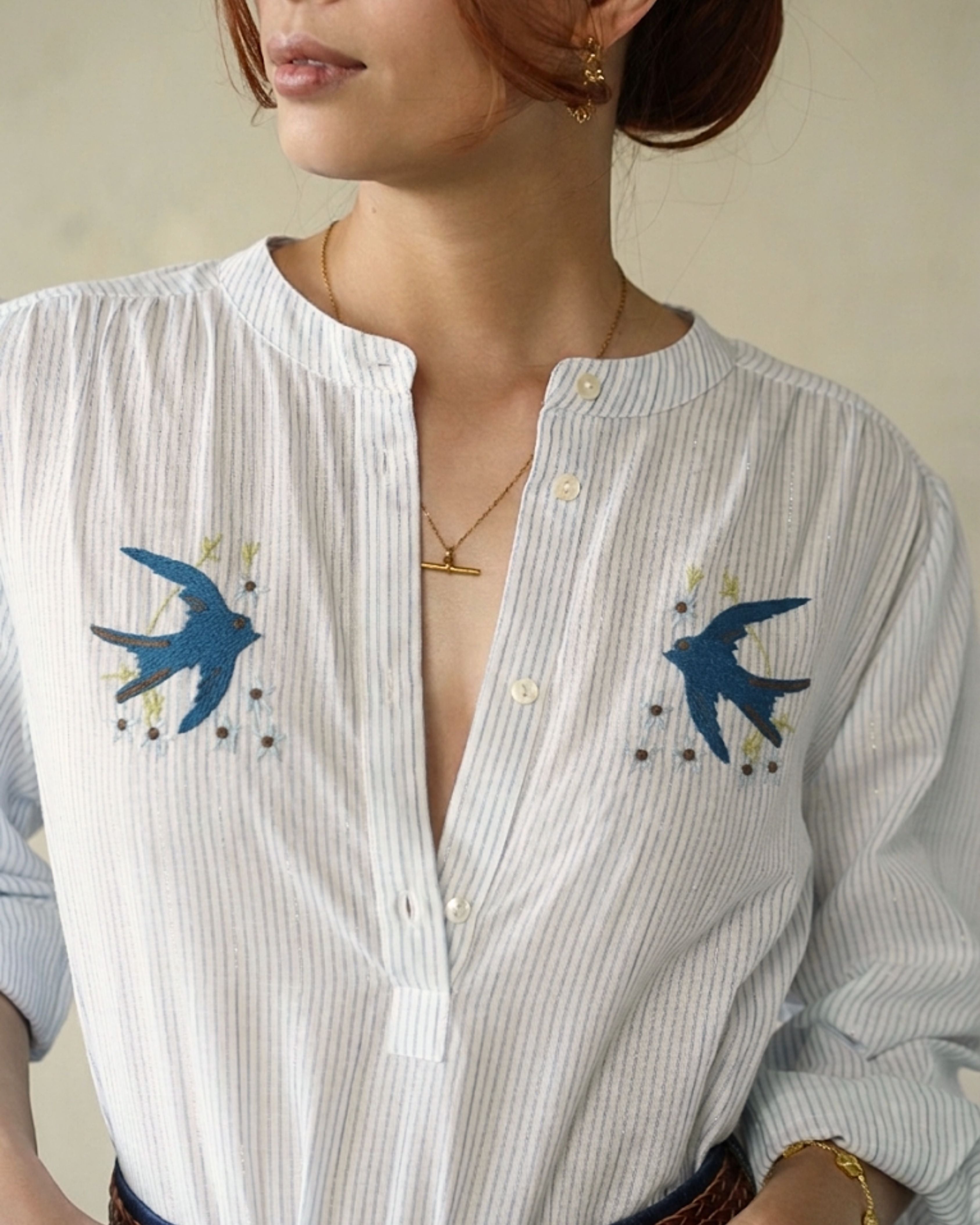 A close-up of a woman wearing a white pinstriped button-up blouse with blue swallow embroidery on both sides of the chest.