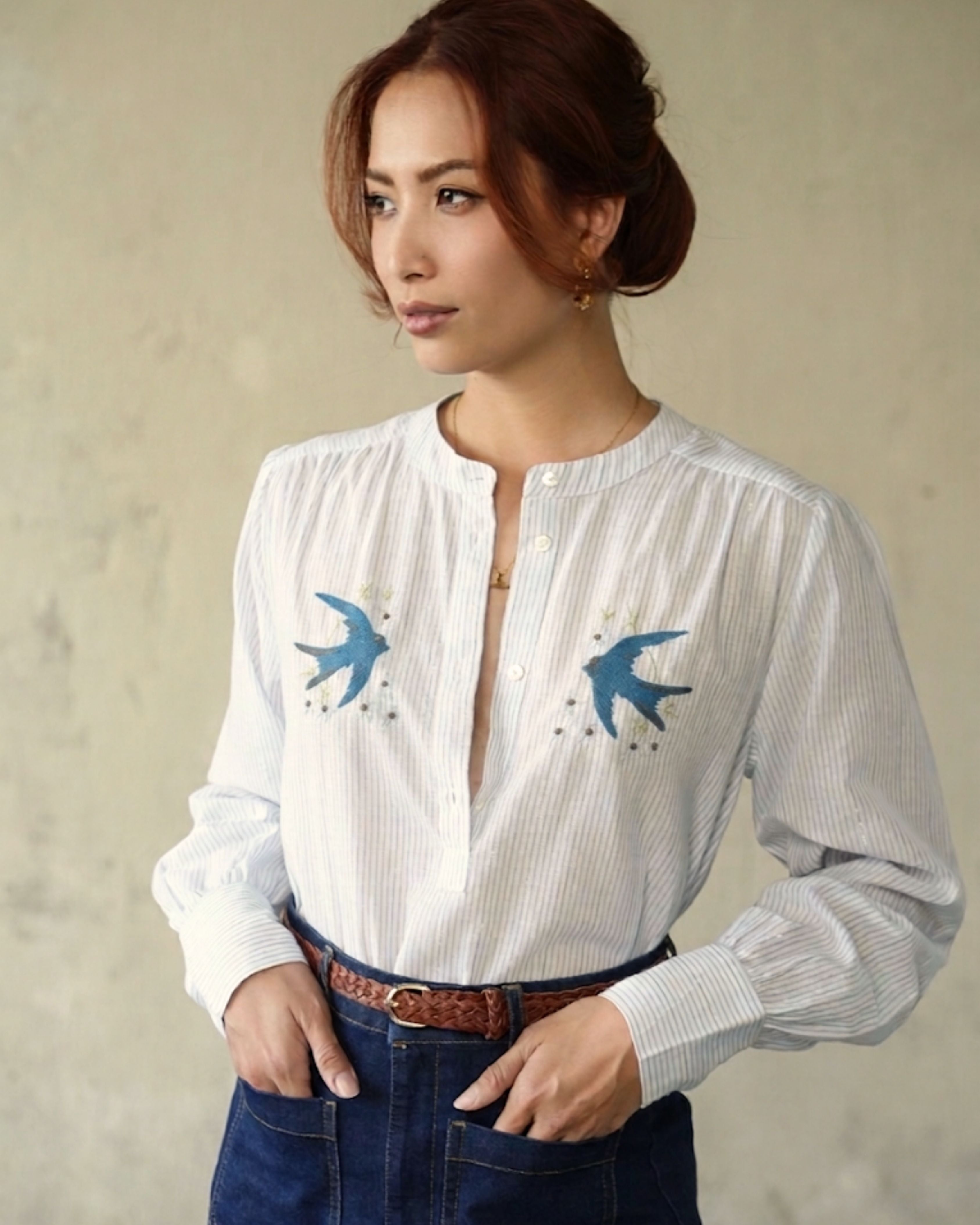 A woman wearing a white long-sleeve button-up blouse with blue swallow embroidery on the chest, paired with dark blue jeans and a brown belt, standing against a textured beige wall.
